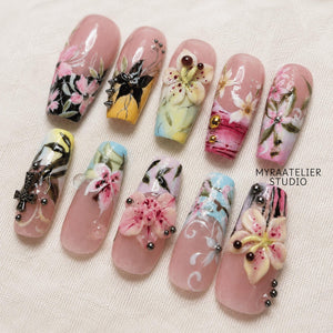 A set of ten press-on nails featuring a nude base with intricate 3D floral designs, hand-painted pink and white flowers, black accents, metallic studs, and cross charms. The text MYRAATELIER STUDIO is printed on the right side.