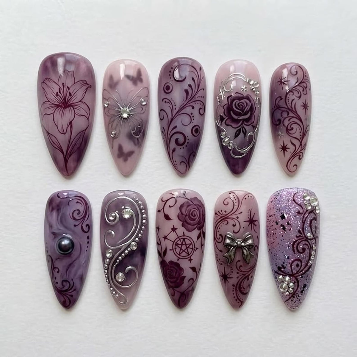 A set of ten almond-shaped press-on nails in shades of purple and mauve. The designs feature dark purple floral patterns, silver metallic swirls, clear rhinestones, a black pearl accent, a silver bow charm, and a pentagram symbol.