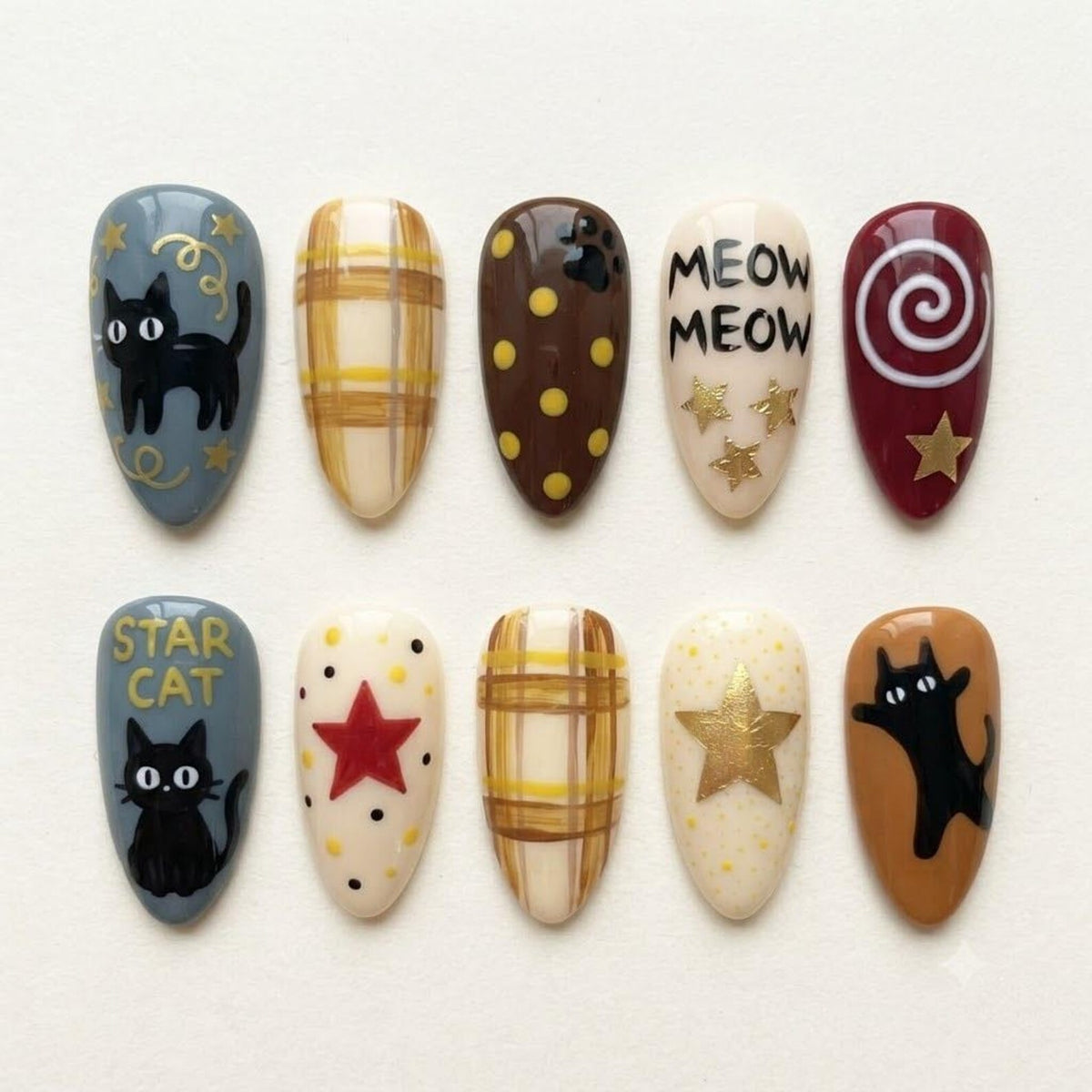 A set of ten almond-shaped press-on nails featuring various designs including black cats, yellow plaid patterns, brown polka dots, gold stars, a red star, and the text MEOW MEOW and STAR CAT painted in black on cream and gray backgrounds.