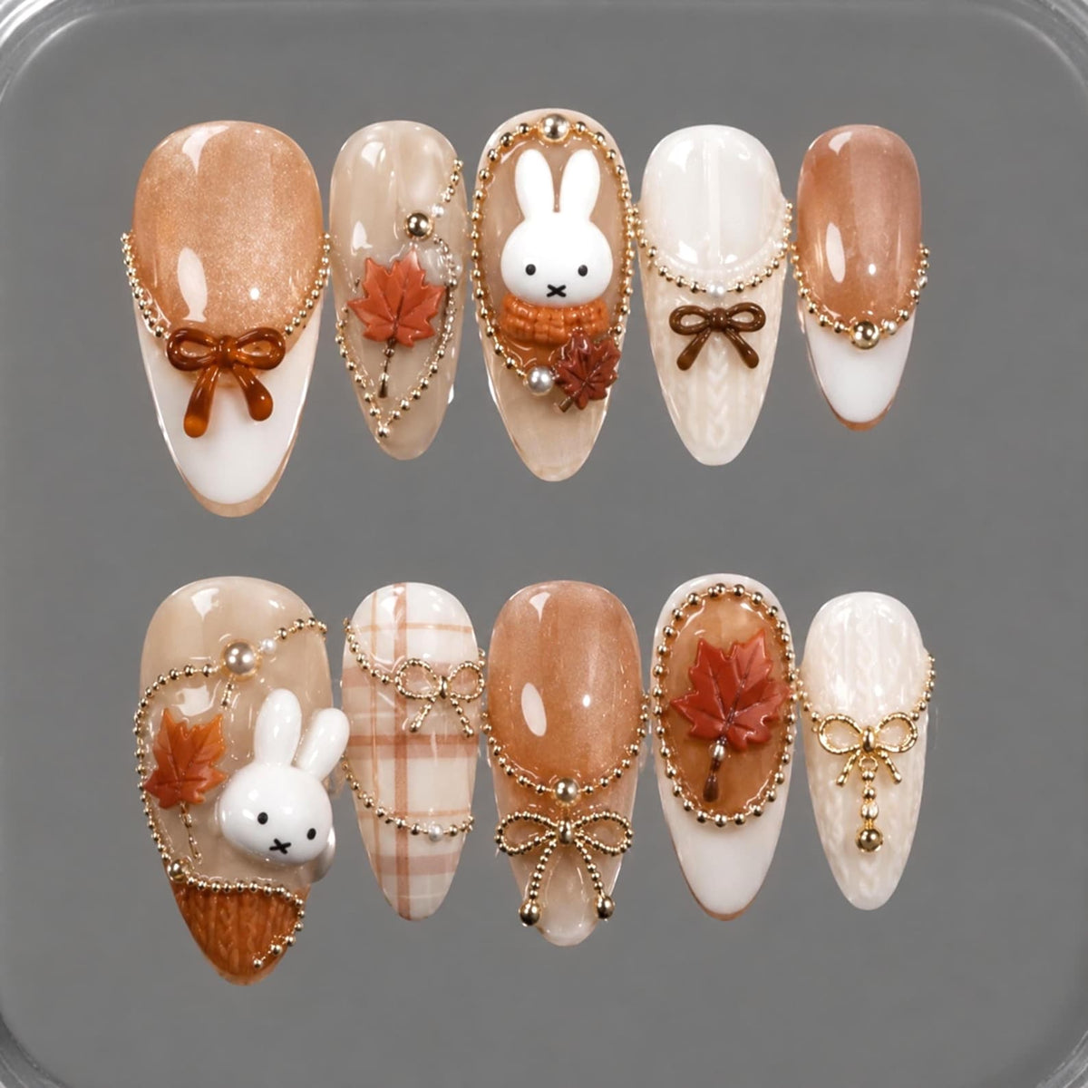 A set of ten almond-shaped press-on nails featuring a fall theme with brown, white, and cream tones. Decorations include 3D white rabbit figures, orange maple leaves, gold beaded chains, and small brown bows on textured surfaces.