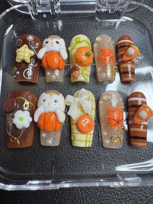 A set of ten press-on nails featuring autumn-themed 3D decorations including orange pumpkins, white cats, maple leaves, plaid patterns, brown stripes, white bows, small flowers, and tiny pearls arranged in a clear plastic storage container.