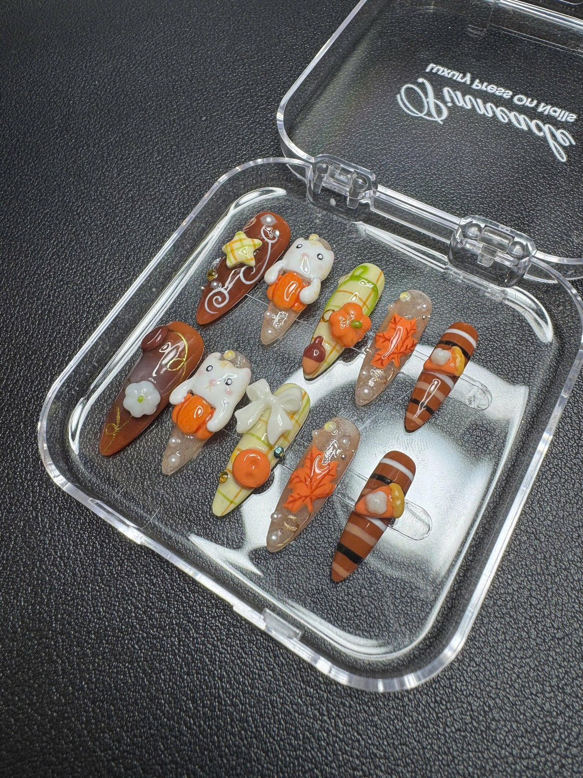 A set of ten press-on nails in a clear plastic case. The nails feature autumn-themed designs including orange pumpkins, white ghosts, brown stripes, maple leaves, and bows in shades of brown, orange, white, and green with gold accents.