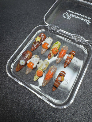 A set of ten press-on nails in a clear plastic case. The nails feature autumn-themed designs including orange pumpkins, white ghosts, brown stripes, maple leaves, and bows in shades of brown, orange, white, and green with gold accents.