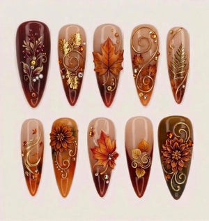 A set of ten almond-shaped press-on nails featuring autumn-themed designs. The nails display deep burgundy, brown, and nude gradients adorned with 3D gold metallic swirls, intricate floral patterns, and detailed orange maple leaves.