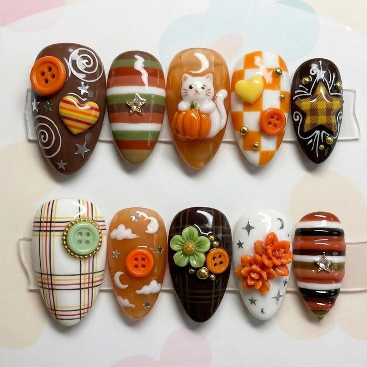 A set of ten press-on nails featuring autumn-themed designs including brown, orange, and white stripes, plaid patterns, a white cat with a pumpkin, orange buttons, hearts, stars, flowers, and succulent-shaped embellishments.