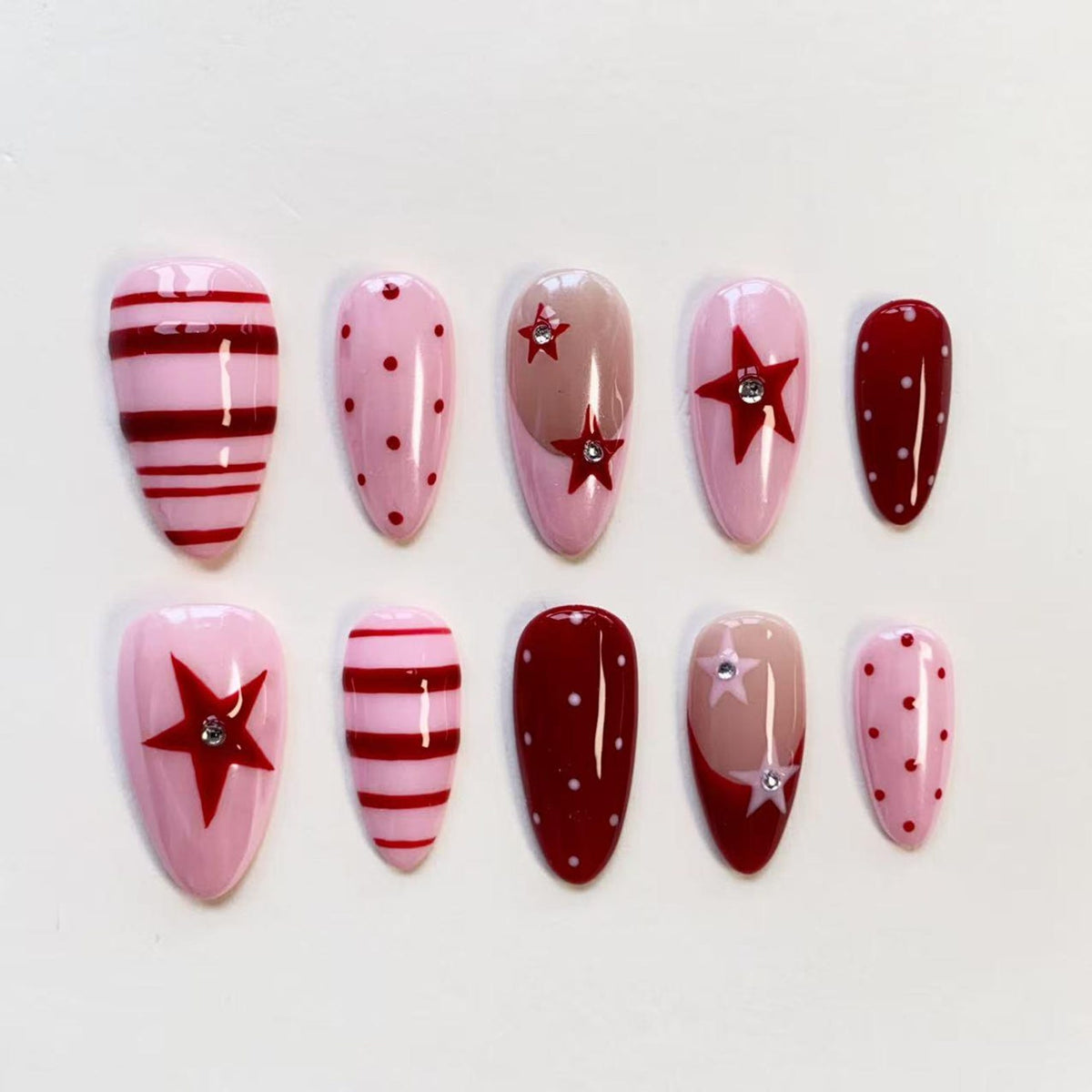 A set of ten almond-shaped press-on nails featuring a color palette of light pink, dark red, and nude. The designs include horizontal stripes, polka dots, and solid red stars, with some stars accented by small, clear rhinestones.