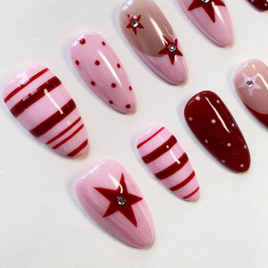 A set of almond-shaped press-on nails featuring a variety of designs including light pink with dark red horizontal stripes, light pink with dark red polka dots, dark red with white polka dots, and light pink with dark red star accents.