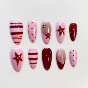 A set of ten almond-shaped press-on nails featuring a color palette of light pink, dark red, and nude. The designs include horizontal stripes, polka dots, and red star motifs accented with small, clear, circular rhinestones.