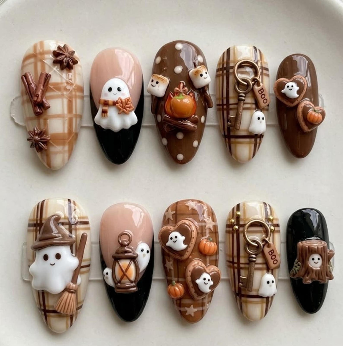 A set of ten press-on nails featuring autumn and Halloween themes. Designs include brown and cream plaid patterns, ghosts, pumpkins, a lantern, a witch hat, a tree stump, and keys with tags labeled BOO, accented with 3D charms.