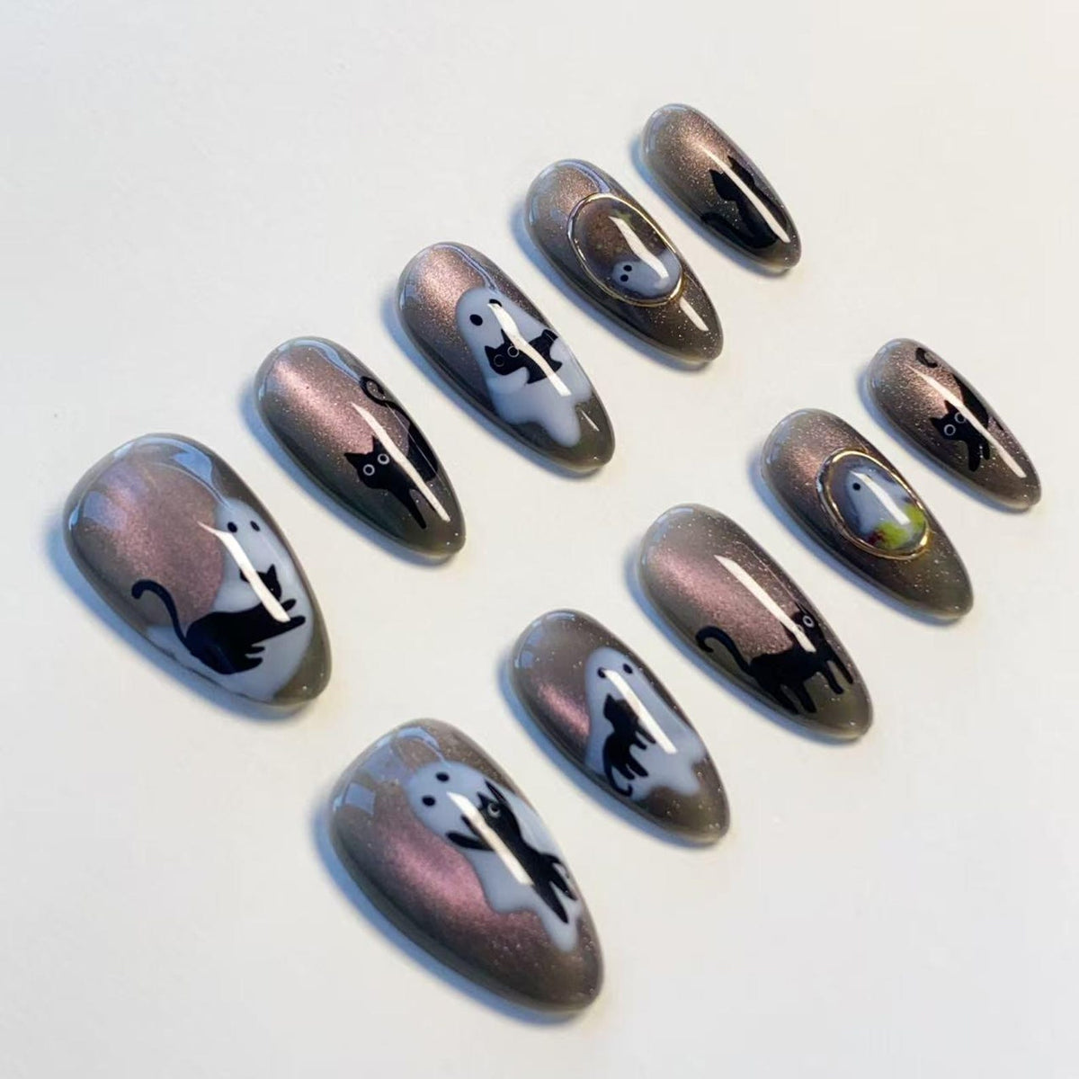 A set of ten almond-shaped press-on nails featuring a shimmering dark gray cat-eye polish base. The designs include black cat silhouettes, white ghost figures, and small gold-toned circular frames on select nails.