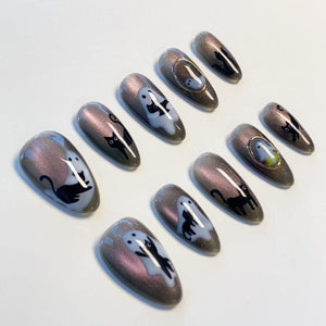 A set of ten almond-shaped press-on nails featuring a shimmering dark gray cat-eye polish base. The designs include black cat silhouettes, white ghost figures, and small gold-toned circular frames on select nails.