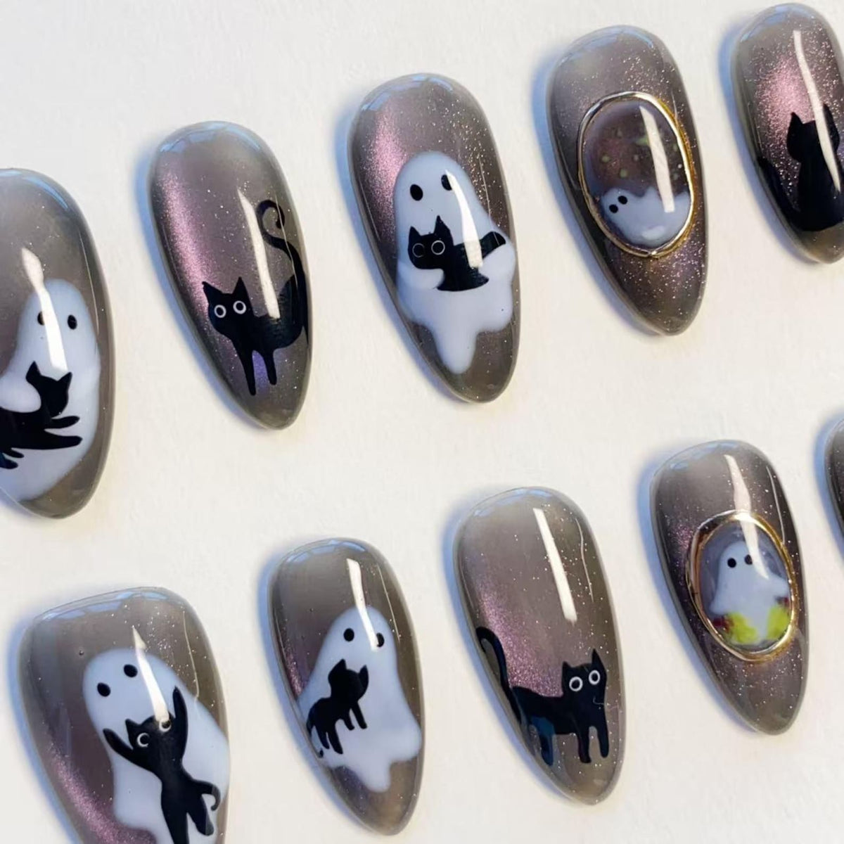 A set of almond-shaped press-on nails featuring a dark gray magnetic shimmer base. The designs include hand-painted black cats, white ghost figures, and small gold-framed accents, creating a cohesive spooky-themed nail art collection.