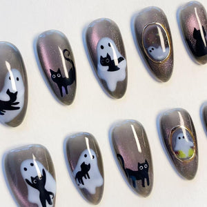 A set of almond-shaped press-on nails featuring a dark gray magnetic shimmer base. The designs include hand-painted black cats, white ghost figures, and small gold-framed accents, creating a cohesive spooky-themed nail art collection.