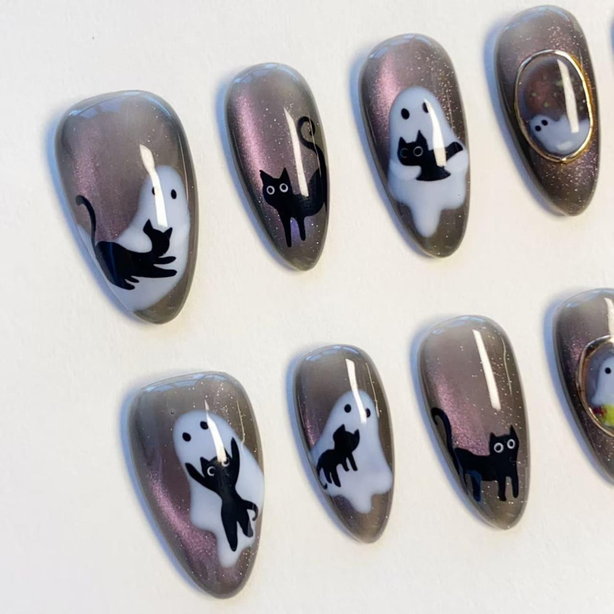 A set of almond-shaped press-on nails featuring a dark grey magnetic cat-eye finish with glitter. The designs include white ghost silhouettes and black cat illustrations, with one nail featuring a small gold-toned oval frame accent.