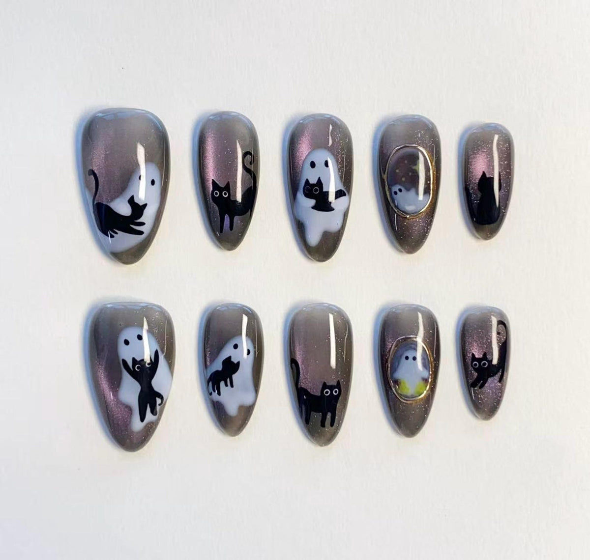 A set of ten almond-shaped press-on nails featuring a dark gray magnetic shimmer base. The designs include white ghost illustrations, black cat silhouettes, and small gold-rimmed oval frames containing tiny ghost motifs.