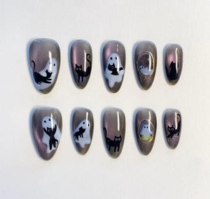 A set of ten almond-shaped press-on nails featuring a dark gray magnetic shimmer base. The designs include white ghost illustrations, black cat silhouettes, and small gold-rimmed oval frames containing tiny ghost motifs.