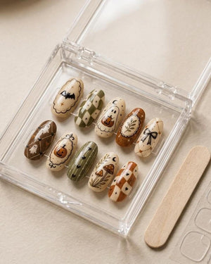 A set of ten oval-shaped press-on nails in a clear plastic storage case. The nails feature autumn-themed designs including ghosts, pumpkins, bats, mushrooms, bows, and checkered patterns in shades of cream, brown, and olive green.