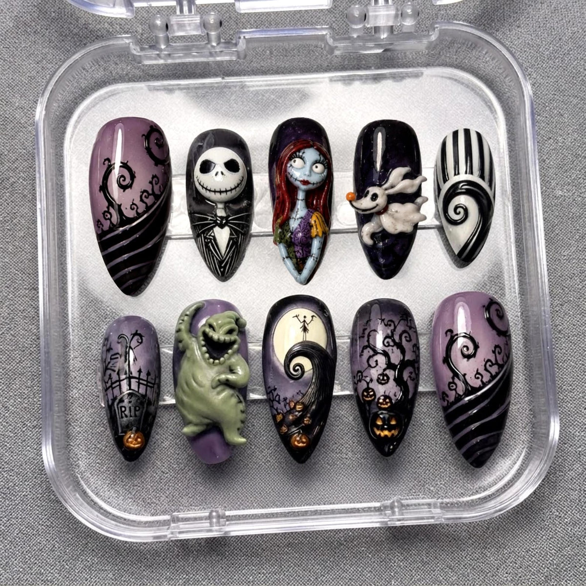 A set of ten almond-shaped press-on nails in a clear plastic case, featuring detailed hand-painted designs inspired by The Nightmare Before Christmas, including characters like Jack Skellington, Sally, Zero, and Oogie Boogie with pumpkins.