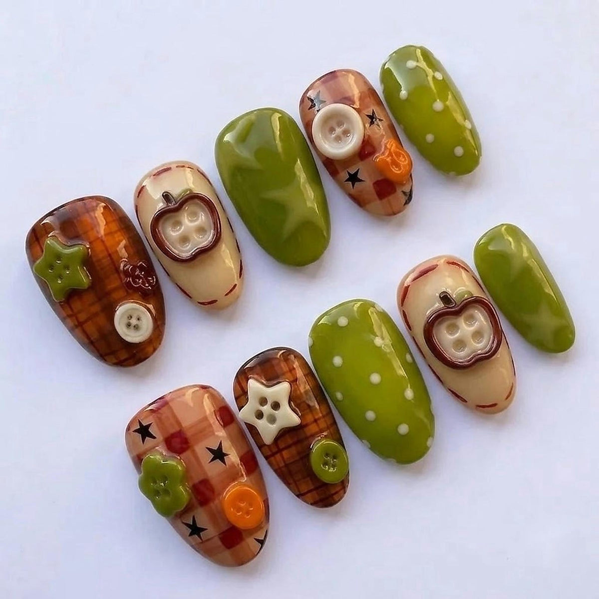 A set of ten press-on nails featuring a variety of designs including brown plaid patterns with black stars, solid olive green surfaces with white polka dots, and cream-colored nails adorned with raised apple-shaped and round buttons.