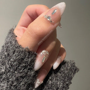 Almond-shaped nails featuring a white and clear gradient base with silver glitter accents. The nail art includes small clear rhinestones, a central iridescent gem, and tiny white pearls arranged in de