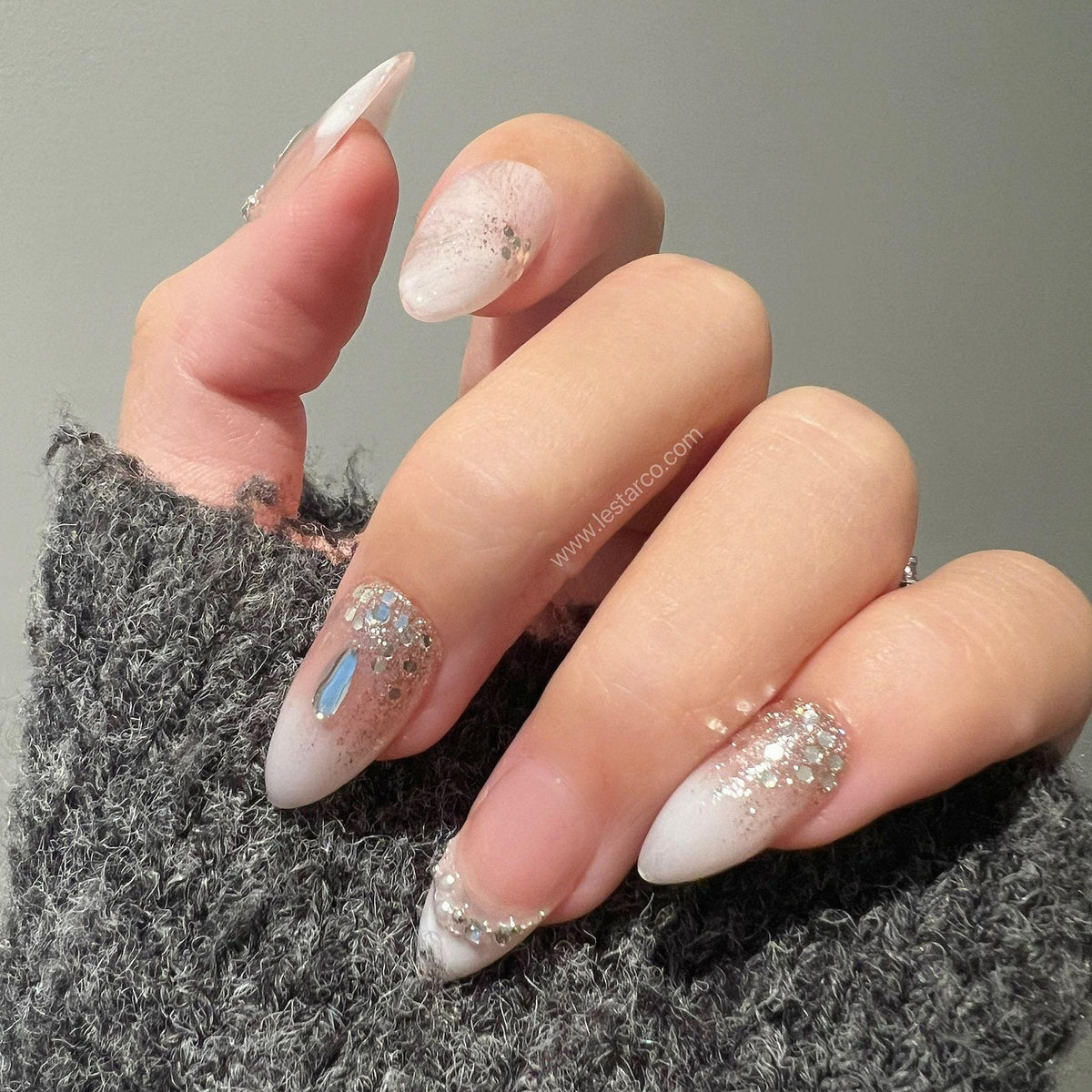 Almond-shaped artificial nails featuring a white ombre gradient base with silver glitter accents and a single blue rhinestone on the ring finger. The nails display a smooth finish and are presented ag
