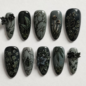 A set of ten almond-shaped press-on nails in shades of charcoal, slate, and black. The designs feature 3D relief elements including black roses, ivy vines, ferns, mushrooms, a beetle, and floral patterns with glossy finishes.