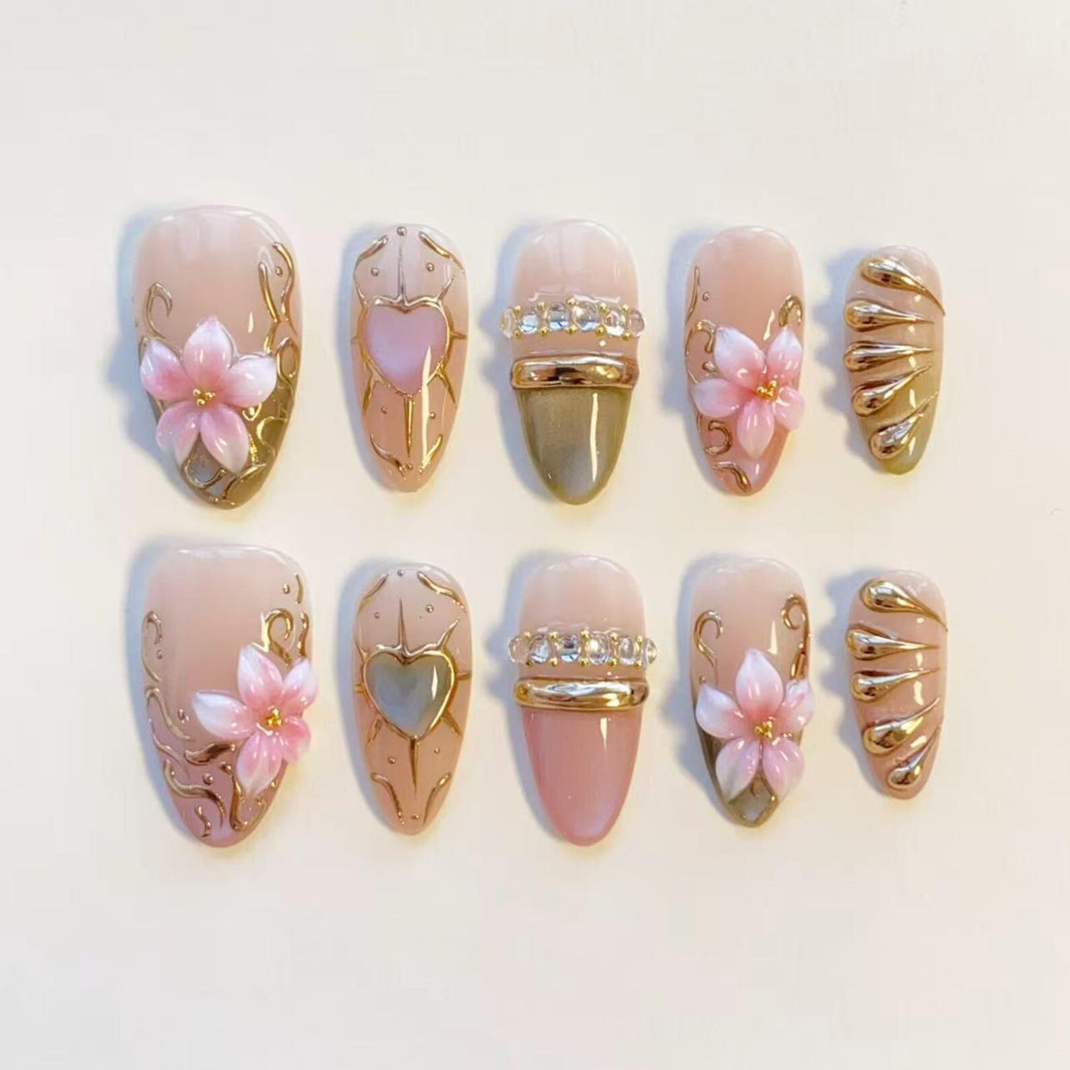 A set of ten press-on nails featuring a nude base with various designs including pink 3D flowers, gold filigree swirls, heart-shaped accents, horizontal gold bands with embedded clear crystals, and raised gold metallic teardrop patterns.