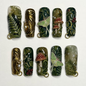 A set of ten rectangular press-on nails featuring intricate 3D designs including green ferns, metallic gold swirls, white pearls, luna moths, red-capped mushrooms, and dark green vines, each with a small gold ring at the base.