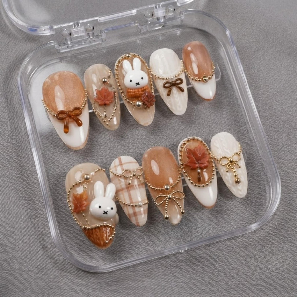 A set of ten almond-shaped press-on nails in a clear plastic case. The nails feature a color palette of white, cream, and brown, decorated with 3D white rabbit figures, brown maple leaves, gold-toned chains, and small bows.