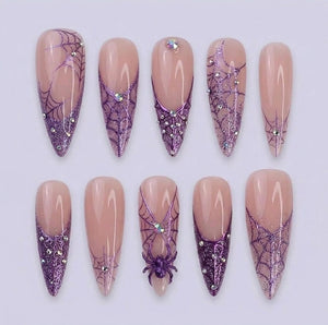 A set of ten almond-shaped press-on nails featuring a nude base with purple glitter spider web designs, intricate web patterns, and small embedded rhinestones. One nail features a three-dimensional purple spider accent.