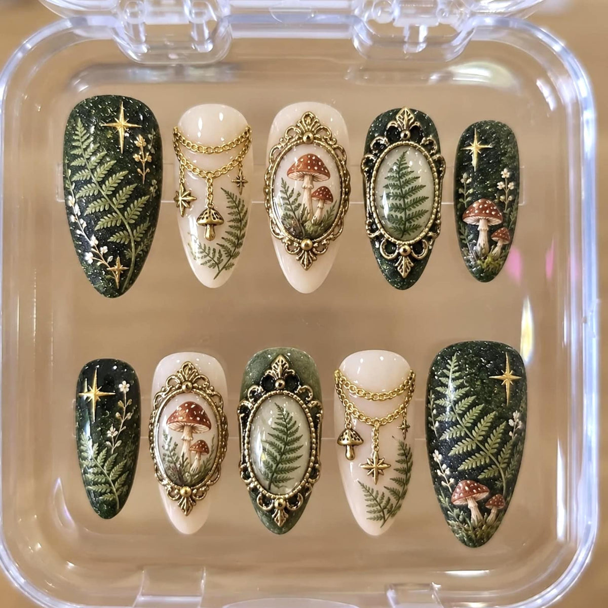 A set of ten almond-shaped press-on nails in a clear plastic case. The designs feature dark green glitter backgrounds with fern and star motifs, cream-colored bases with gold chain charms, and ornate gold frames enclosing mushroom or fern art.