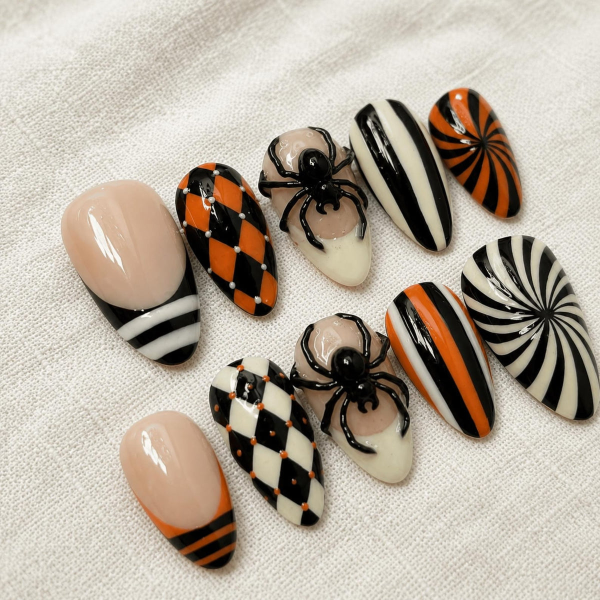 A set of ten press-on nails featuring almond-shaped tips with various designs including black and orange harlequin patterns, black and white vertical stripes, orange and black swirls, and two nails adorned with raised black 3D spider figures.