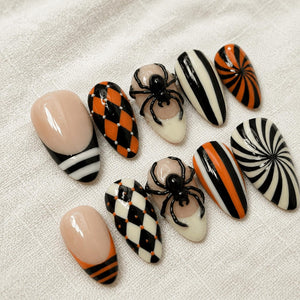 A set of ten press-on nails featuring almond-shaped tips with various designs including black and orange harlequin patterns, black and white vertical stripes, orange and black swirls, and two nails adorned with raised black 3D spider figures.