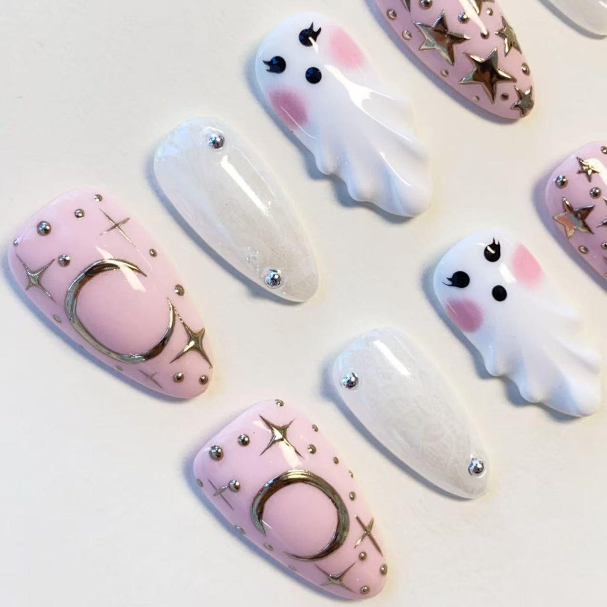 A set of press-on nails featuring a light pink base with gold crescent moon and star accents, white ghost figures with pink cheeks, and translucent white nails with silver studs. The designs include metallic gold stars and dots.