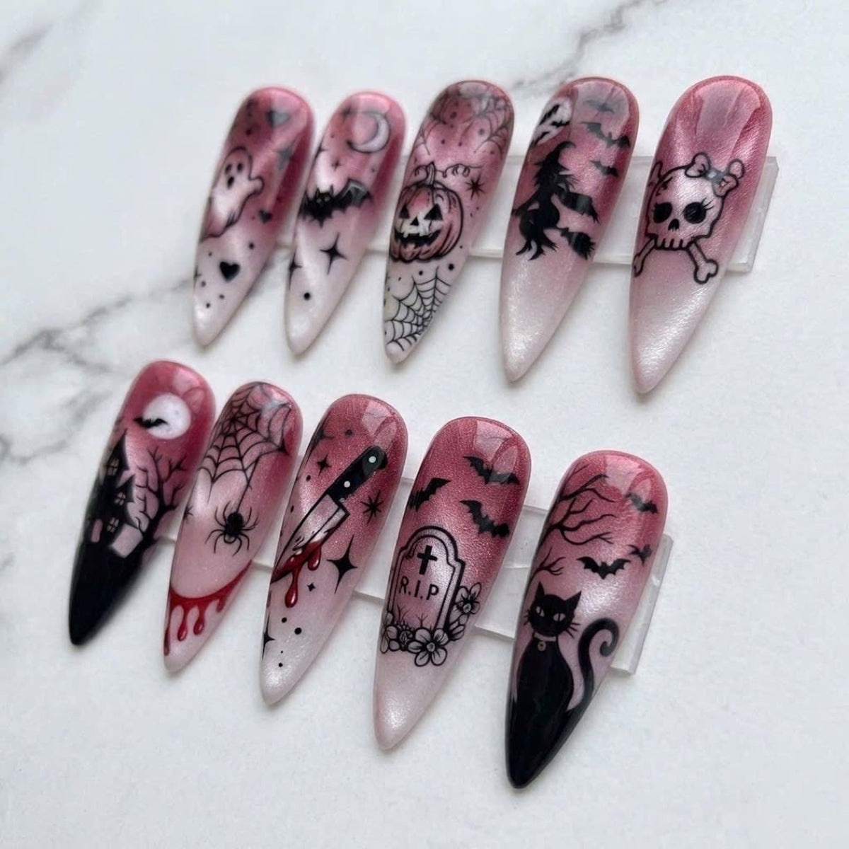 A set of ten stiletto-shaped press-on nails featuring a pink and white gradient base with black Halloween-themed designs including a ghost, bat, pumpkin, witch, skull, haunted house, spider web, knife, tombstone with RIP, and cat.