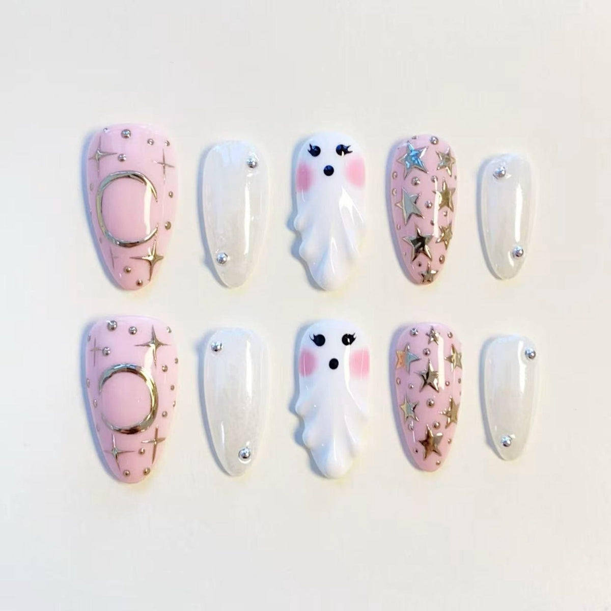 A set of ten press-on nails featuring a light pink base with silver moon and star accents, white nails with small silver studs, and white ghost-shaped nails with pink cheeks and black eyes, arranged in two rows of five.