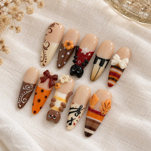 A set of ten almond-shaped press-on nails featuring various designs including brown swirls, polka dots, a ladybug, piano keys, a white bow, orange stripes, a small cat figure, musical notes, and orange autumn leaves.