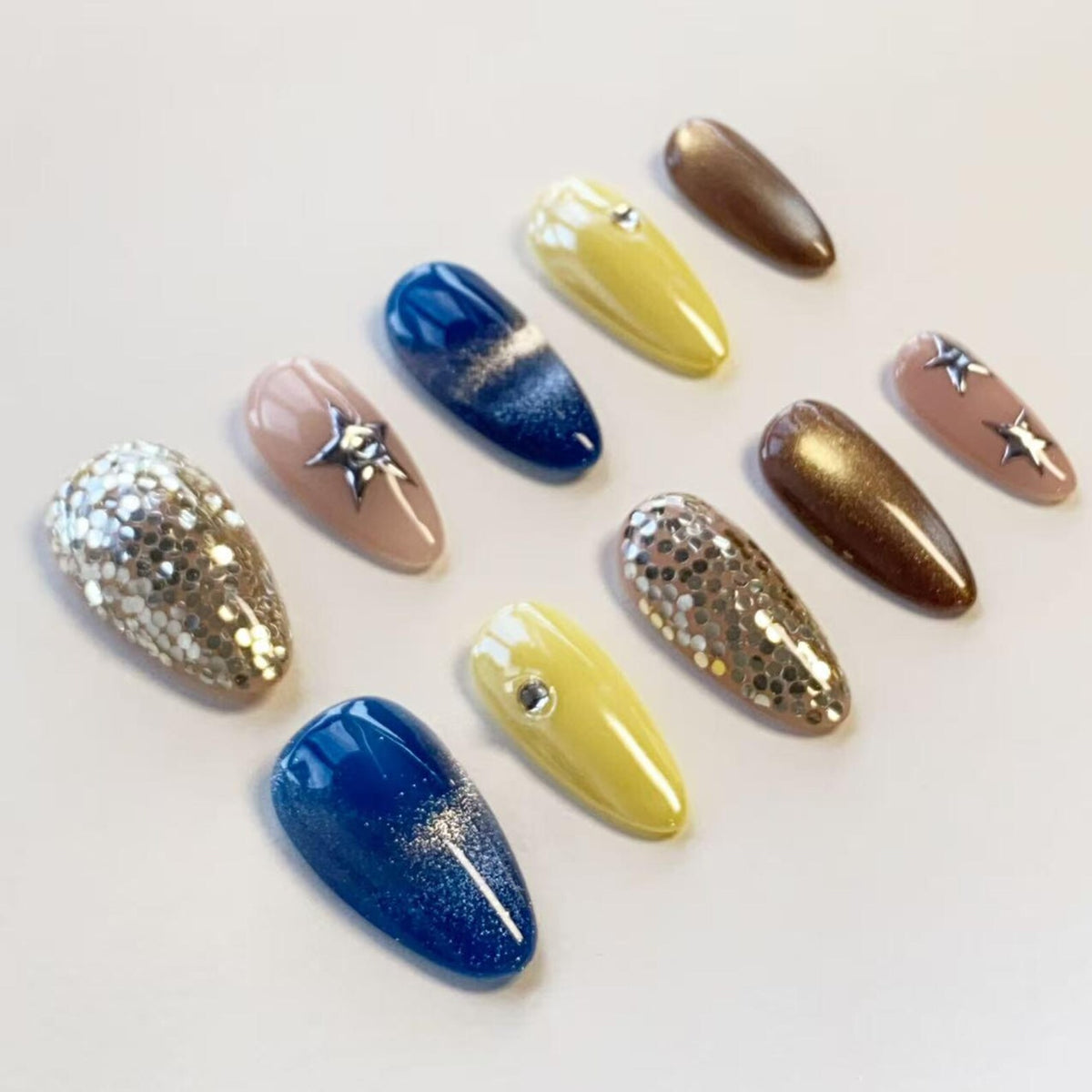 A set of ten almond-shaped press-on nails featuring various designs including solid yellow, deep blue with a glitter gradient, metallic bronze, nude with silver star accents, and full silver hexagonal glitter coverage.