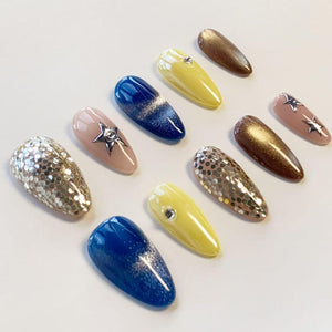 A set of ten almond-shaped press-on nails featuring various designs including solid yellow with a single rhinestone, deep blue with silver glitter gradients, nude with silver star charms, gold glitter, and large silver sequins.