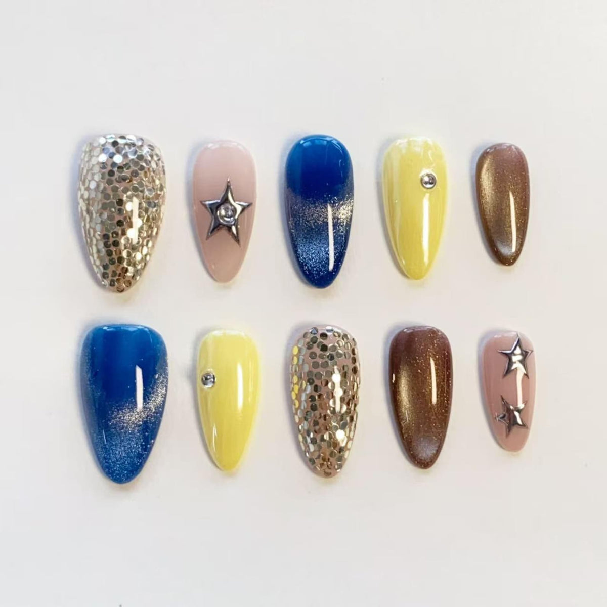 A set of ten almond-shaped press-on nails featuring various designs including silver glitter, solid yellow with a single rhinestone, deep blue with silver shimmer, metallic bronze, light pink with silver star charms, and clear rhinestones.