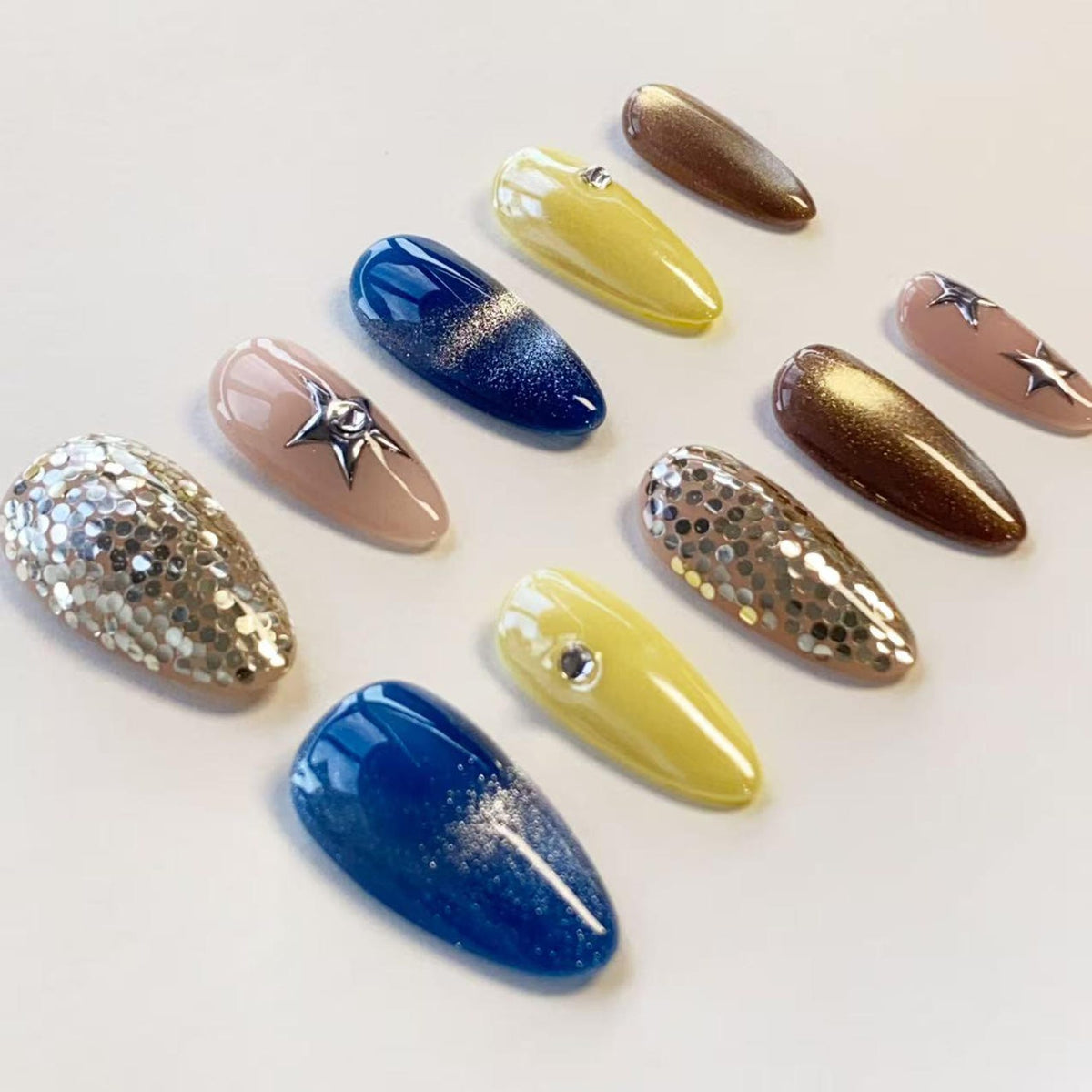 A set of ten almond-shaped press-on nails featuring various designs including solid yellow, deep blue with silver glitter gradients, brown metallic, nude with silver star charms, and full coverage silver sequin embellishments.