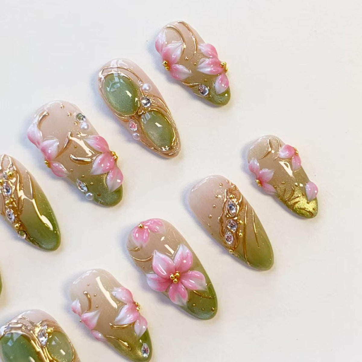 A set of almond-shaped press-on nails featuring a gradient of sage green and nude beige. The designs include hand-painted pink cherry blossom flowers, gold metallic line accents, small white pearls, and clear rhinestones.