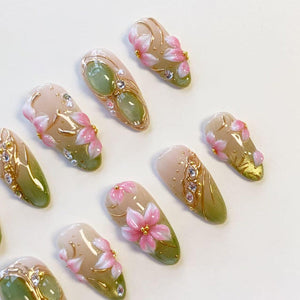 A set of almond-shaped press-on nails featuring a gradient of sage green and nude beige. The designs include hand-painted pink cherry blossom flowers, gold metallic line accents, small white pearls, and clear rhinestones.
