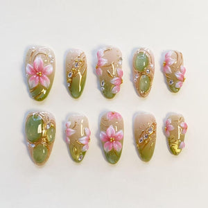 A set of ten press-on nails featuring a gradient of soft pink and sage green. The designs include raised 3D pink flower petals, translucent green oval accents, gold metallic line detailing, and small clear rhinestones on each nail.