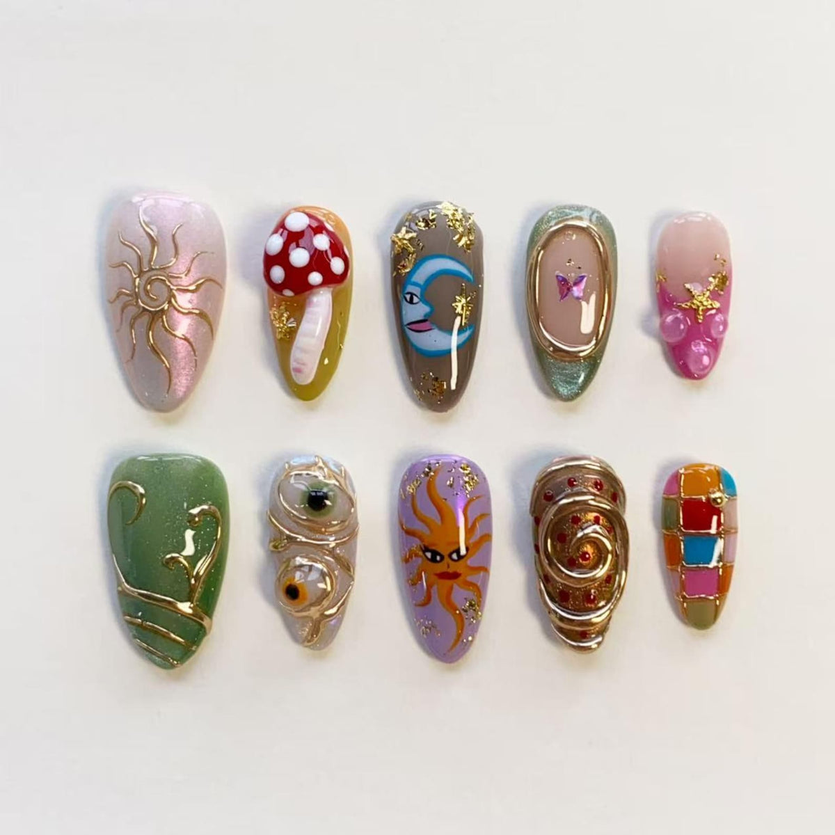 A set of ten press-on nails featuring diverse designs including a gold sun, a 3D red and white mushroom, a blue crescent moon, a butterfly, pink accents with gold stars, green swirls, two eyes, a sun face, gold spirals, and a colorful grid.