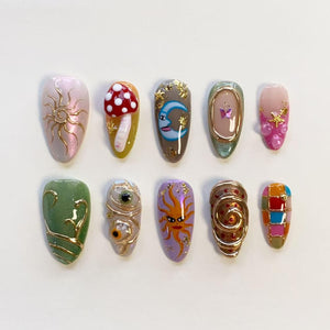 A set of ten press-on nails featuring diverse designs including a gold sun, a 3D red and white mushroom, a blue crescent moon, a butterfly, pink accents with gold stars, green swirls, two eyes, a sun face, gold spirals, and a colorful grid.