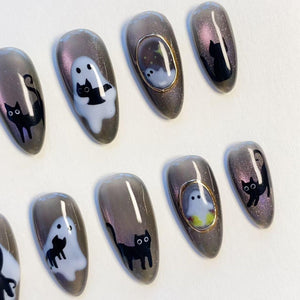 A set of almond-shaped press-on nails featuring a dark gray cat-eye magnetic polish base. The designs include black cat silhouettes, white ghost figures, and small oval frames with gold metallic borders containing tiny ghost illustrations.