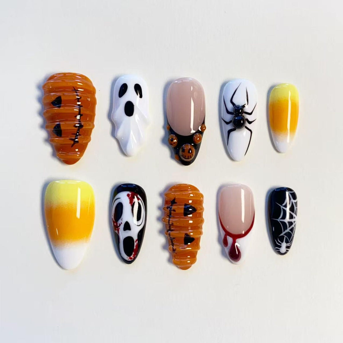 A set of ten press-on nails featuring Halloween designs including orange pumpkins, white ghosts, a black spider on white, candy corn gradients, a horror movie mask with red blood splatters, and a black nail with a white spider web pattern.