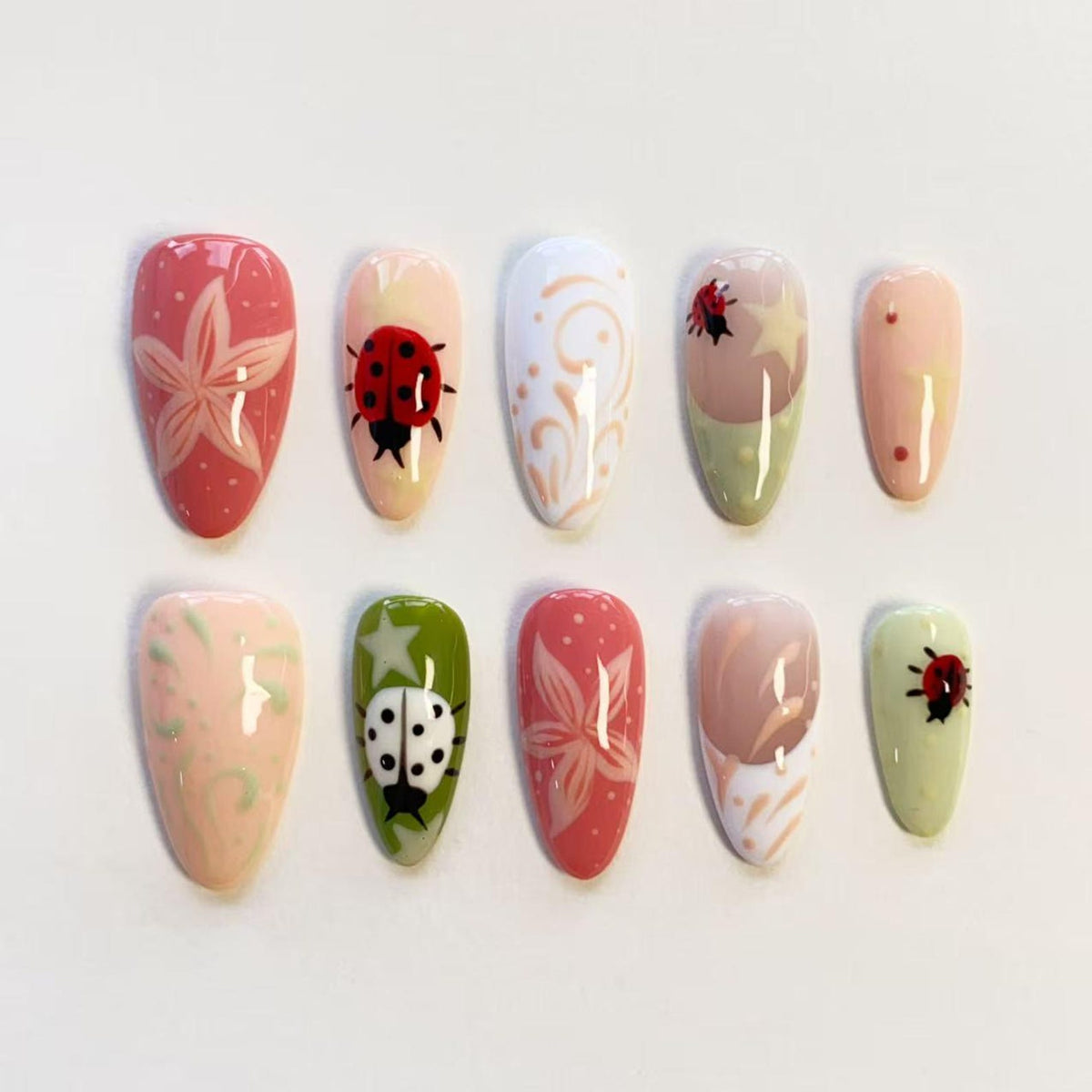 A set of ten almond-shaped press-on nails featuring various designs including red ladybugs, white star patterns, pink floral motifs, and swirling lines in shades of pink, green, white, and beige with a glossy finish on each nail.