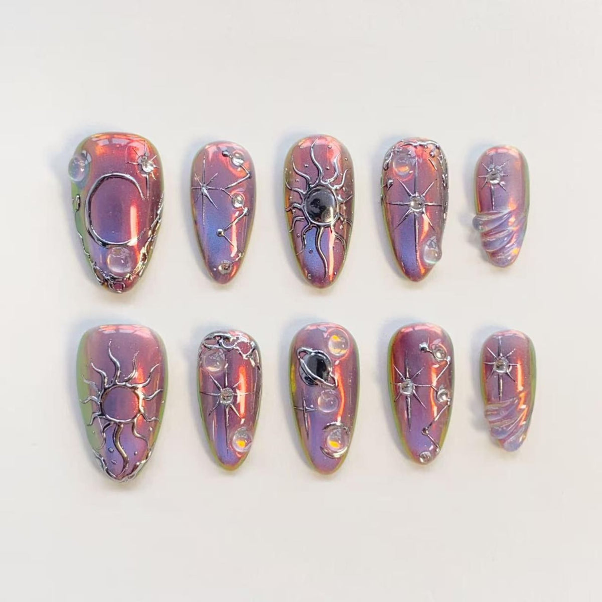 A set of ten almond-shaped press-on nails with a metallic, iridescent purple and pink finish. Each nail features raised silver celestial designs including suns, moons, stars, and planets, accented with small, clear, spherical embellishments.